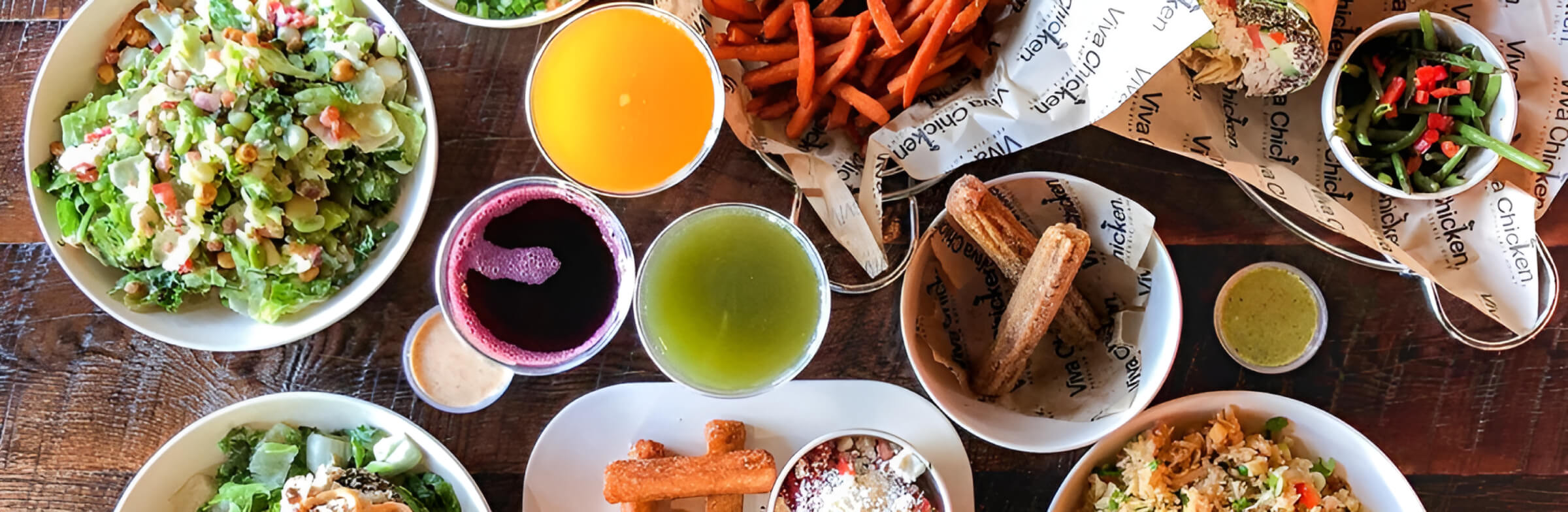 A wooden table topped with various dishes, including salads, sweet potato fries, bread, sauces, juice drinks, and bowls of rice and vegetables, arranged for a colorful meal.