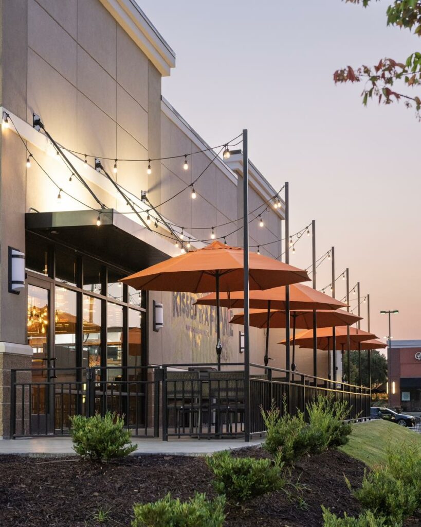 Viva Chicken restaurant patio at dusk with orange umbrellas, string lights, outdoor seating, and large windows. Small bushes line the foreground under a clear evening sky.