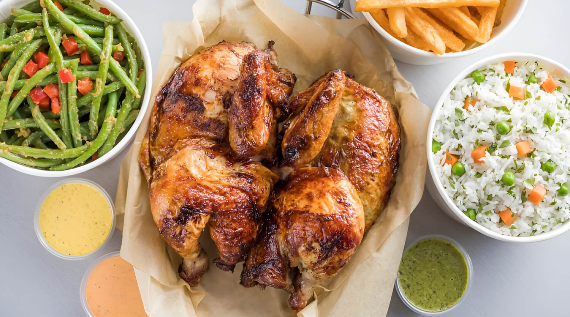A roasted chicken sits on parchment paper, surrounded by bowls of green beans with tomatoes, seasoned fries, rice with peas and diced vegetables, and small cups of various dipping sauces.
