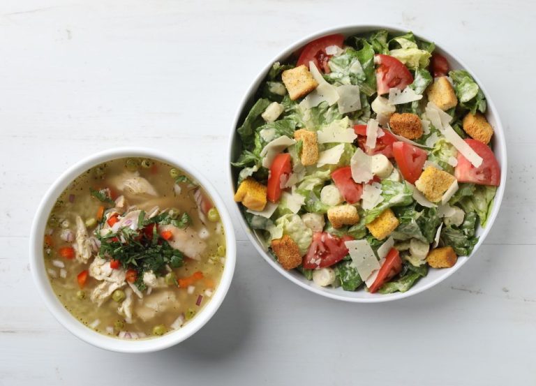 A bowl of chicken soup with herbs beside a bowl of Caesar salad topped with croutons, parmesan cheese, and tomato slices on a white surface.